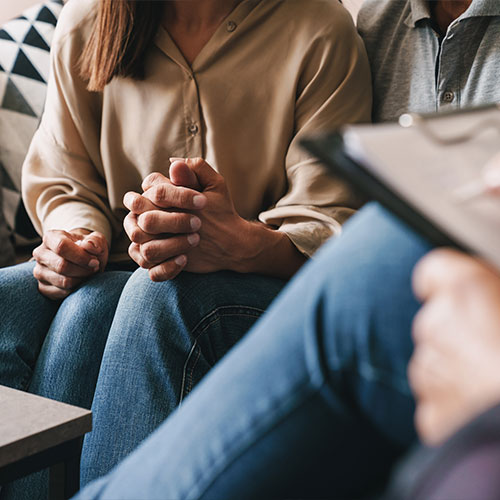 Anonymes Paar sitzt händchenhaltend im Hintergrund. Im Vordergrund sitzt eine dritte anonyme Person mit einem Klemmbrett auf den überschlagenen Beinen.