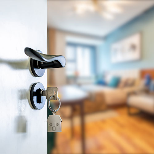 Geöffnete Tür mit Schlüssel im Türschloss, an dem ein Haus-Anhänger befestigt ist. Im Hintergrund sieht man verschwommen einen Raum.