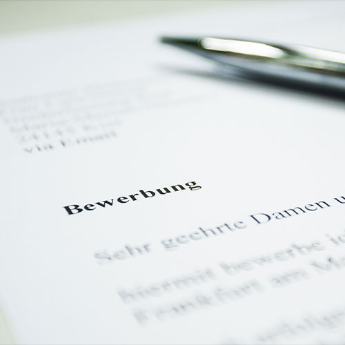 Blatt Papier auf dem "Bewerbung" geschrieben steht. An der oberen rechten Kante liegt ein Kugelschreiber auf dem Papier.