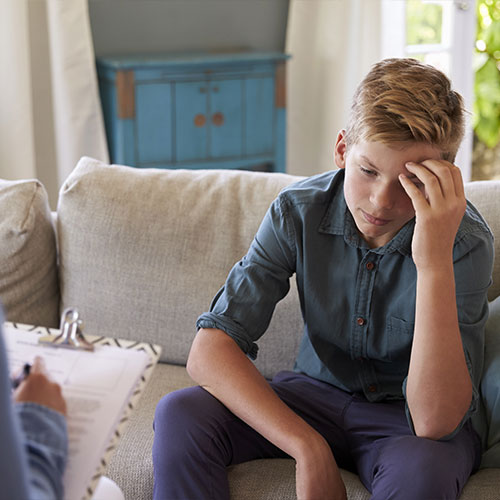 Jugendlicher sitzt eingesunken und mit seiner linken Hand an seiner Stirn auf einem Sofa. Ihm gegenüber sitzt eine anonyme Person, die auf einem Klemmbrett Notizen macht.