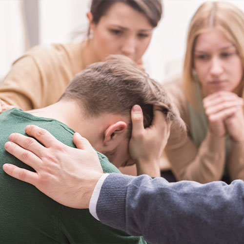 Junger Mann mit Kopf in seinen Händen und drei weitere Personen, die ihm zuhören und Trost spenden.