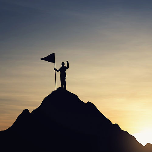 Silhouette eines Berges vor dem Morgenhimmel mit einem Menschen auf der Spitze, der eine Fahne auf die Bergspitze stellt und winkt.